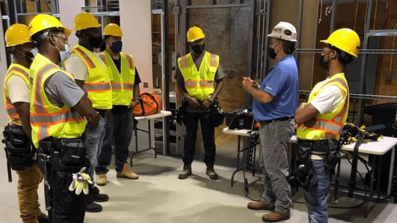 A group of men dressed in construction gear stand a semi circle in an indoor job site as a foreman gives instructions.