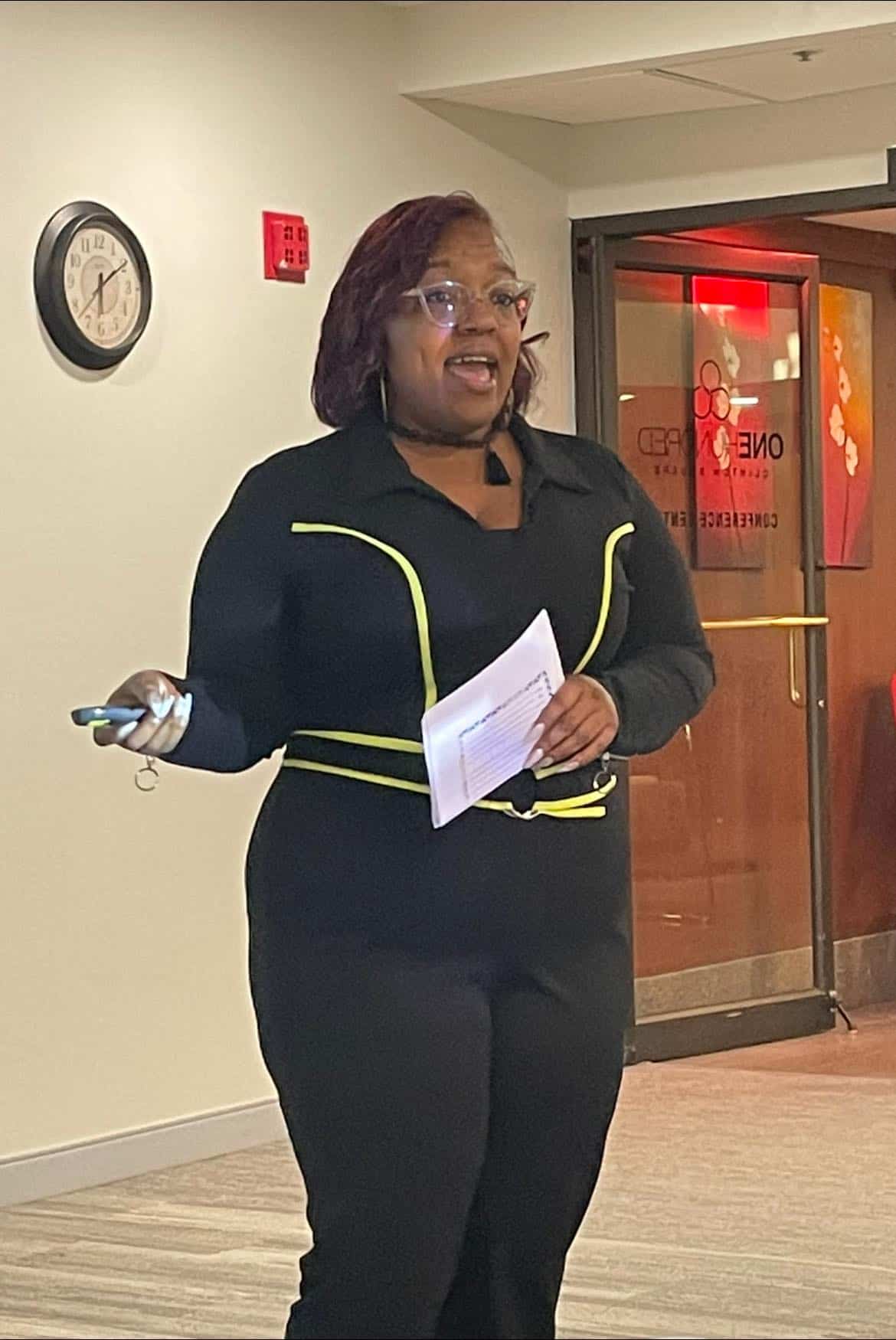 Executive Director Sheena Solomon addresses the room wearing a black and yellow jumpsuit while holding some documents..