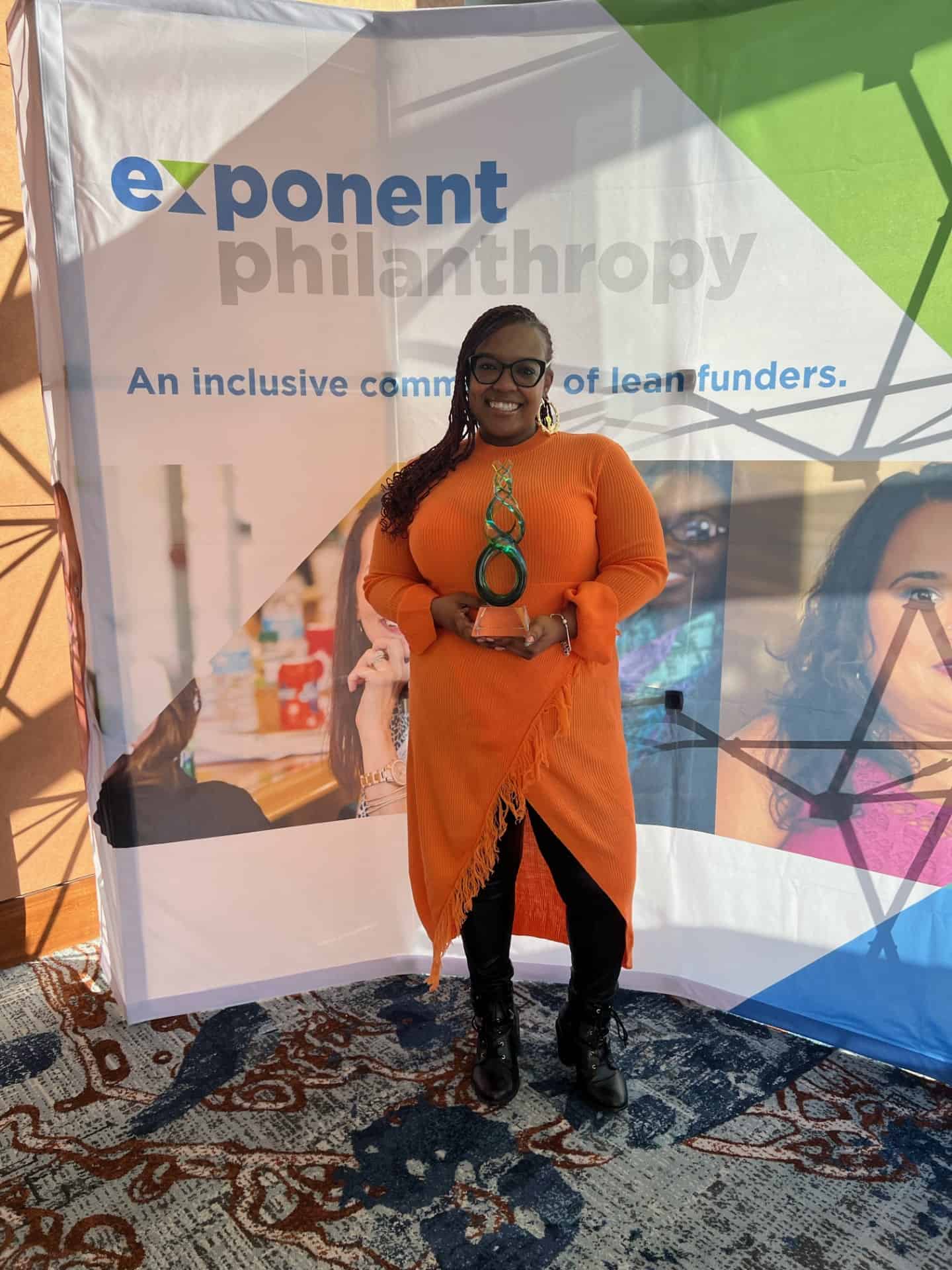 Gifford Executive Director Sheena Solomon smiles and poses with her award, She is wearing a tight orange dress and braided hair.