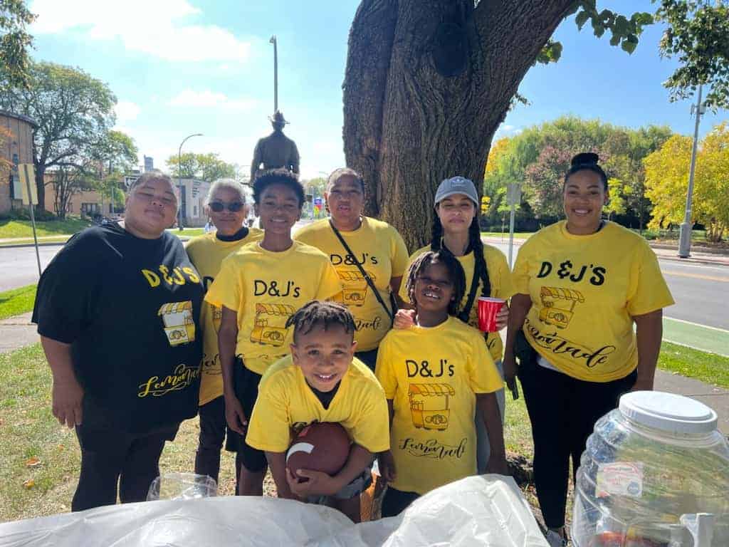 A group of kids and adults wearing yellow shirts that say "D&J's Lemonade" pose next to a table.