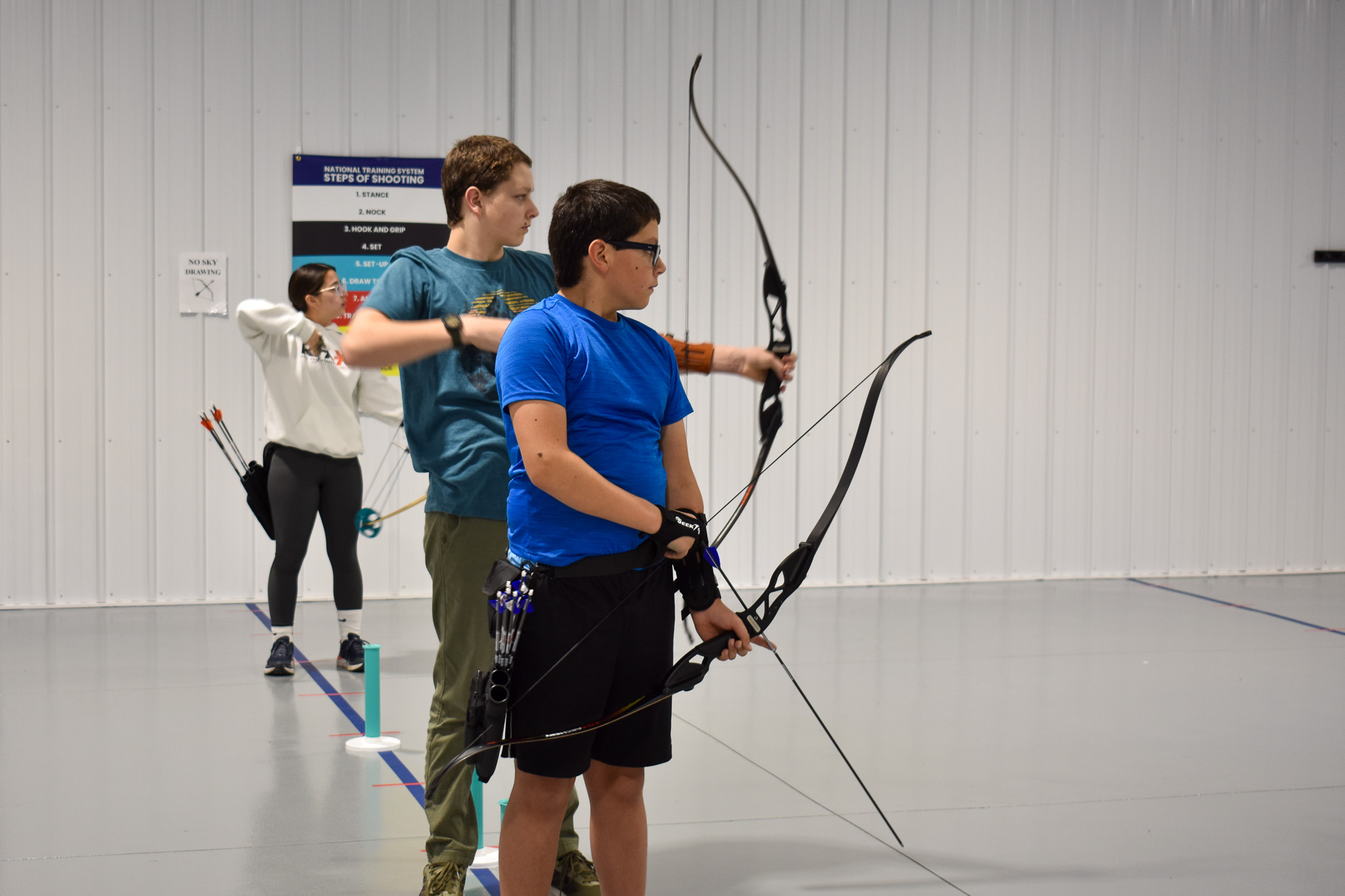 A boy in middle school with a royal blue shirt on in profile holding a bow and arrow getting ready to take a shot.