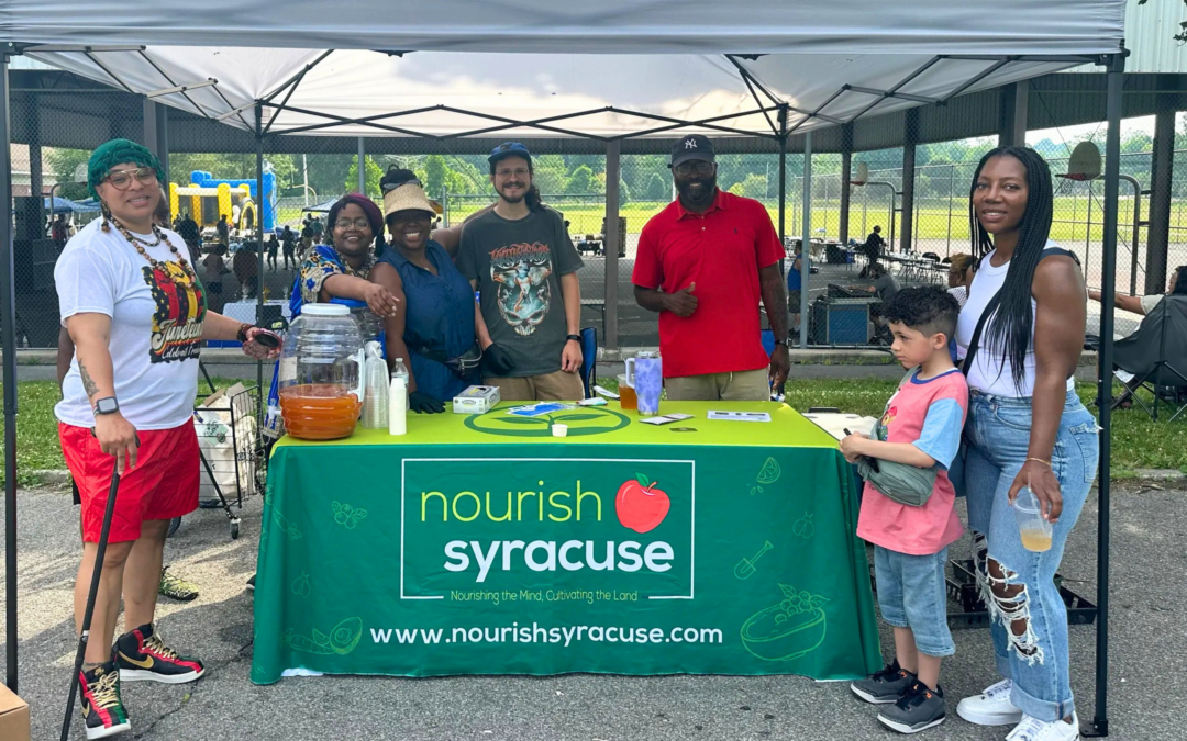 A group of adults and one child standing at a outdoor tented booth on a sunny day for Nourish Syracuse - all smiling. The table under the tent has a green branded Nourish Syracuse table cloth on it.