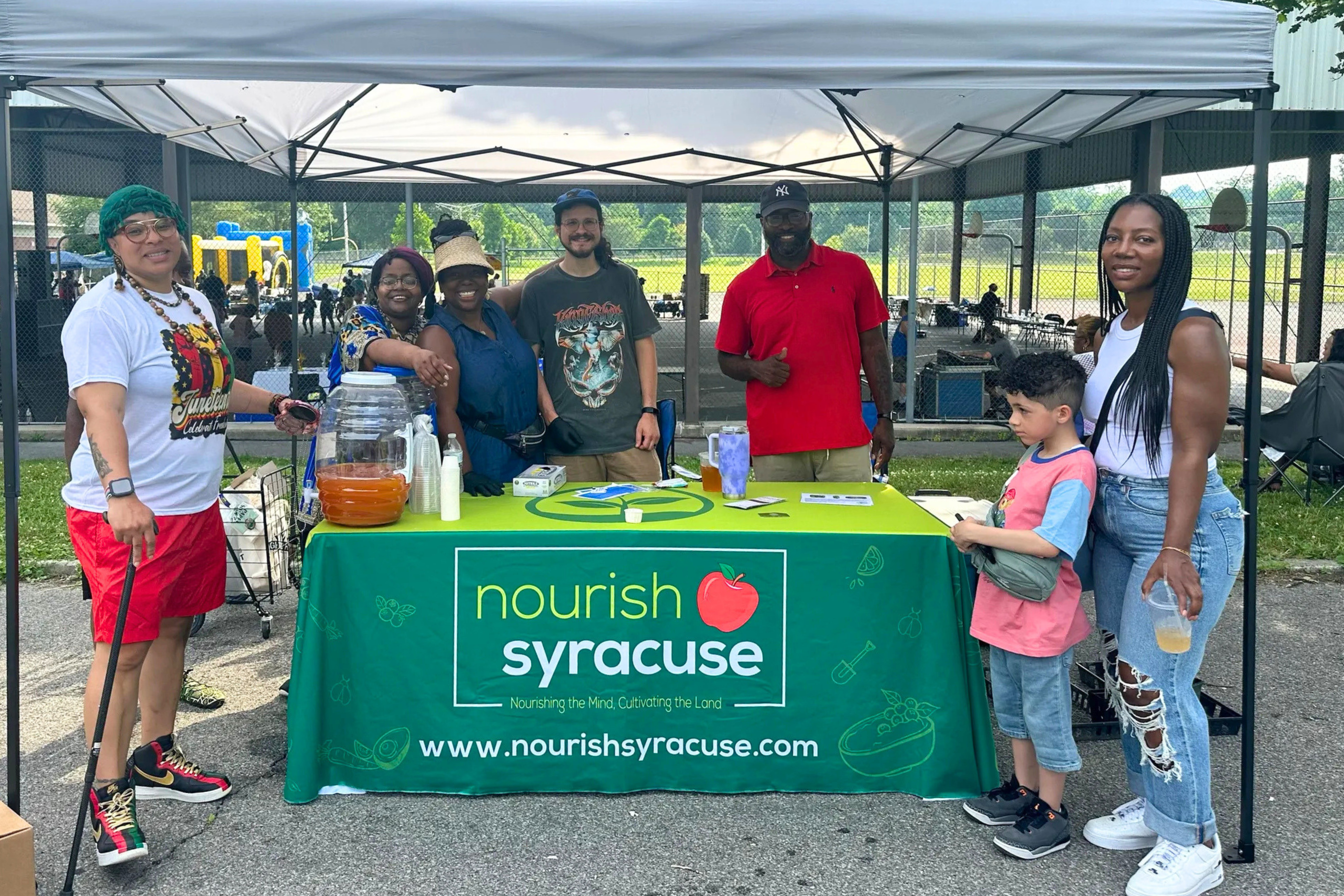 A group of adults and one child standing at a outdoor tented booth on a sunny day for Nourish Syracuse - all smiling. The table under the tent has a green branded Nourish Syracuse table cloth on it.