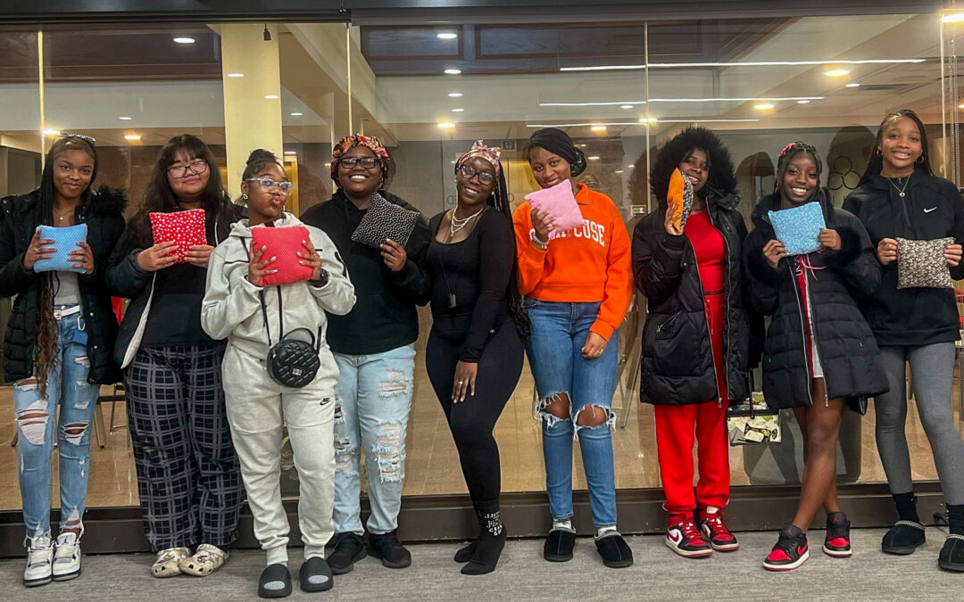 A group of around 10 African American teen girls standing next to eachother with pillows they've made, smiling.