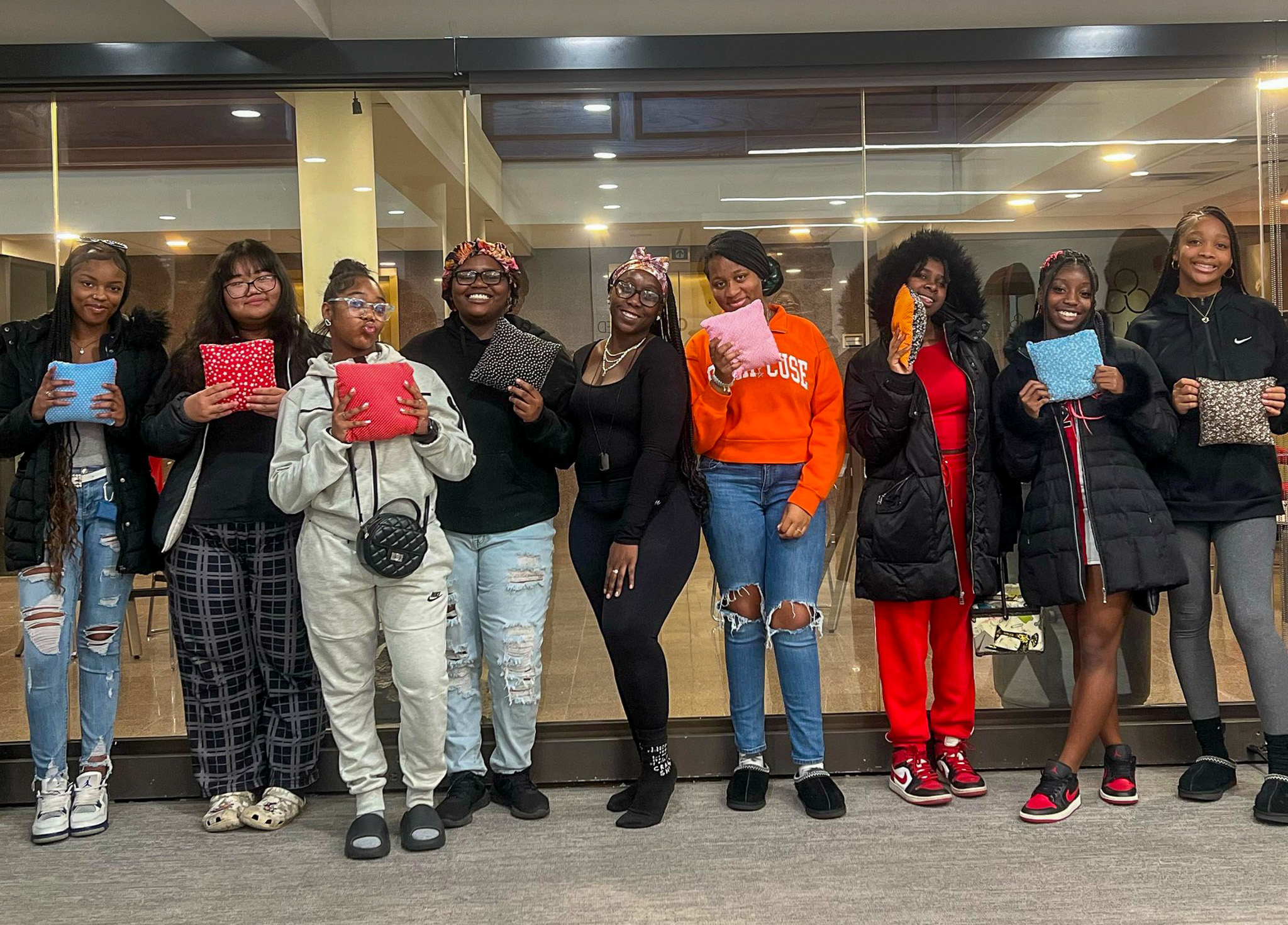 A group of around 10 African American teen girls standing next to eachother with pillows they've made, smiling.