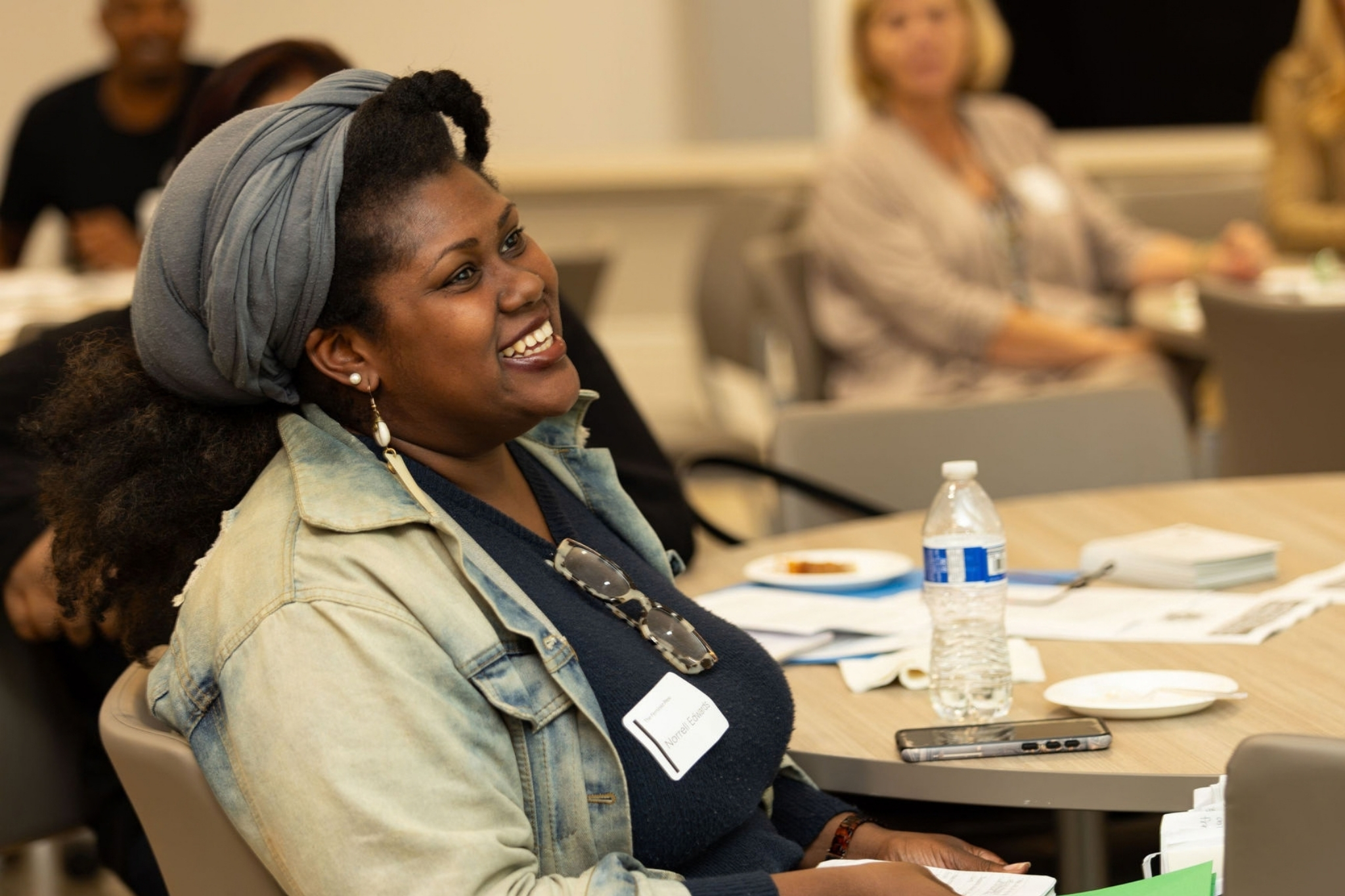 NTL participant smiling while sitting at a table at the kick off sessio of NTL. She is an African American woman wearing a head scarf and a distressed jean jacket.